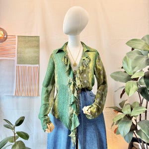 Op de afbeelding: Een groene blouse met lange mouwen, een ruche aan de voorzijde, een kraag en een subtiel ton-sur-ton patroon. Het kledingstuk wordt getoond op een etalagepop en gecombineerd met een blauwe spijkerbroek. Een goudkleurige hangerketting is zichtbaar.