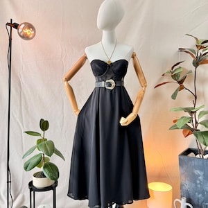 Puede incluir: Un vestido midi negro sin tirantes con un corpiño de encaje y una falda fluida. La prenda se ajusta a la cintura con un cinturón negro que tiene una hebilla plateada prominente. Un collar con colgante dorado descansa sobre el escote.