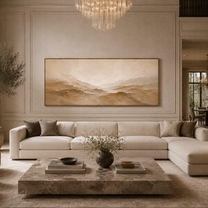 May include: A large horizontal landscape painting in shades of beige and brown hangs above a long cream-colored sectional sofa. The room features a low square stone coffee table, two rounded armchairs, and a crystal chandelier overhead.
