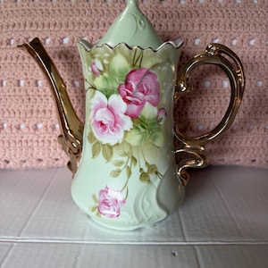 May include: A vintage porcelain teapot with a light green base, adorned with pink roses and gold accents. The teapot features a curved handle, a spout, and a lid with a gold finial. The floral design adds a touch of elegance to this decorative piece.