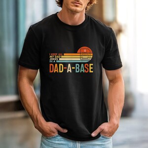 May include: A black short-sleeved t-shirt featuring a graphic with a retro-style sunset design in orange, yellow, and blue. The text reads I KEEP ALL MY DAD JOKES IN A DAD-A-BASE, accompanied by a line-art illustration of a floppy disk icon.
