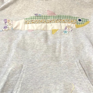 May include: A light gray hooded sweatshirt featuring a large fish applique on the chest. The fish is constructed from various fabric patches including green gingham, floral prints, and solid cream, with a dark blue eye and striped dorsal fin.