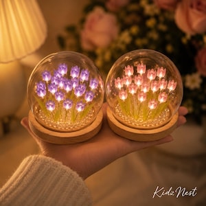 Peut inclure: Deux globes en verre avec des arrangements de tulipes illuminées. L'un présente des tulipes violettes, l'autre roses. Chaque globe repose sur une base en bois, avec des perles à la base des fleurs. Les globes sont tenus dans une main.