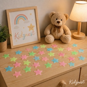 Może przedstawiać: Oprawiony wydruk z tęczą i gwiazdami, pluszowy miś i kolorowe dekoracje w kształcie gwiazd. Na wydruku napis "Kidznest". Gwiazdy są niebieskie, różowe i zielone. Widoczna jest również lampa i mała roślina.
