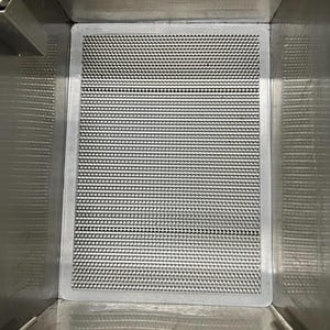 May include: A rectangular, stainless steel mesh filter with a gray border, set inside a stainless steel container. The mesh has a grid-like pattern. The container has a reflective surface.