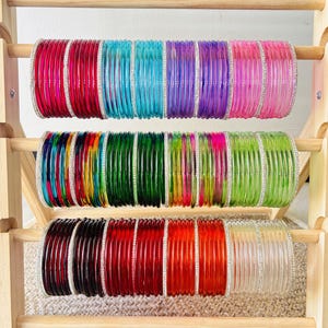 May include: A wooden display stand holding three horizontal rows of colorful bangles. The bangles feature various solid and translucent hues including red, blue, purple, pink, green, orange, and clear, with thin silver-toned accents between them.