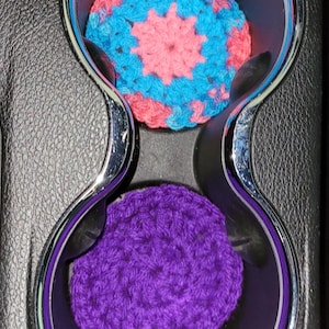 May include: Two crocheted car coasters. One coaster is blue with a pink starburst design. The other coaster is solid purple. The coasters are placed in a car's cup holders, which are black with chrome accents.