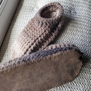 May include: A pair of ankle-high slippers featuring a textured, light brown crocheted upper with a ribbed pattern. The soles are made of a dark brown, smooth, leather-like material that is stitched to the crocheted body with visible thread.