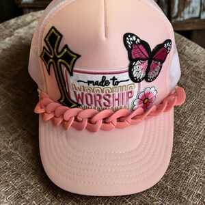 Peut inclure: Casquette rose clair de type trucker avec un dos en filet blanc. Elle présente une croix dorée et noire, un papillon et l'inscription "made to WORSHIP". Une bordure rose en forme de chaîne orne la visière.