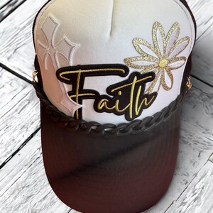 Peut inclure: Casquette de camionneur blanche et marron avec le mot "Faith" en lettres dorées. La casquette présente une marguerite dorée à paillettes et un motif de feuille décoratif. La visière est marron et l'arrière est en mesh marron.