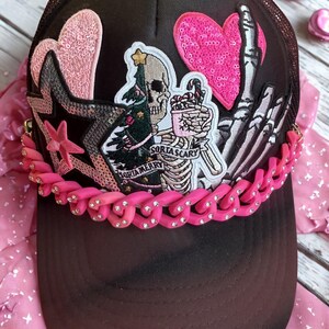 Peut inclure: Casquette de camionneur noire avec un dos en filet et une visière incurvée. L'avant présente des écussons d'un squelette avec un sapin de Noël, des cœurs roses et une étoile. Une chaîne rose avec des strass orne la visière. Le texte sur l'écusson se lit "Sorta Scary" et "Sorta Merry."