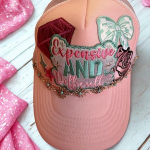 Puede incluir: Gorra trucker rosa claro con parte trasera de malla y visera curva. La gorra presenta bordados, incluyendo un diamante, un lazo y una rosa. El texto "Expensive and Difficult" está bordado en la parte delantera.