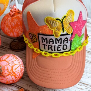 Peut inclure: Casquette de camionneur pêche avec un panneau avant blanc. Le chapeau présente un papillon jaune, des paillettes en forme d'étoile roses et vertes, et le texte "MAMA TRIED" sur un patch blanc. Une chaîne jaune décore le bord.