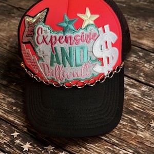 Puede incluir: Gorra de camionero negra con panel frontal rojo y parte trasera de malla marrón. La gorra presenta el texto "Expensive AND Difficult" en un diseño bordado colorido, junto con adornos de estrellas y signos de dólar. Una cadena plateada está unida a la visera.