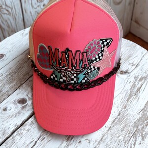 Peut inclure: Casquette de camionneur rose vif avec le mot "MAMA" dans un design coloré. Le chapeau comporte une chaîne noire sur le bord et un dos en filet. Le design comprend une étoile, une fleur et un drapeau à damier.