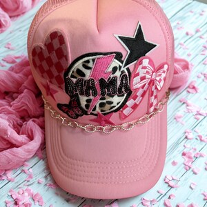 Peut inclure: Casquette de camionneur rose clair avec un dos en filet et une visière incurvée. La casquette présente des écussons brodés, dont un cœur, une étoile, un nœud et le mot "MAMA" dans un motif léopard. Une chaîne dorée est attachée à la visière.