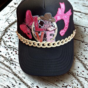 Peut inclure: Casquette de camionneur noire avec un dos en filet et une visière incurvée. L'avant présente un papillon rose, le mot "LOVE" et un motif de cow-girl. Une chaîne crème traverse la visière.