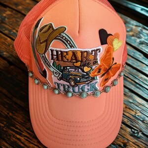 Peut inclure: Casquette de camionneur corail avec un dos en filet. L'avant présente un motif brodé avec un chapeau de cowboy, un fer à cheval, un papillon et les mots "Heart Like a Truck". Orné de pierres de couleur turquoise.