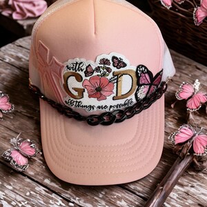 Peut inclure: Casquette rose avec un dos en filet blanc et une visière incurvée. Le chapeau présente un écusson brodé avec les mots "with GOD all things are possible" et des motifs floraux et papillons. Un détail de chaîne noire orne l'avant.