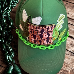 Peut inclure: Casquette de camionneur verte avec un dos en filet et une visière incurvée. La casquette présente des écussons brodés, dont une étoile, un papillon et le texte "SOMEBODY'S FINE ASS BABY MAMA". Une chaîne vert fluo est attachée à la casquette.