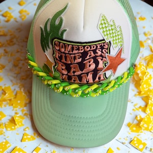 Peut inclure: Casquette de camionneur verte et blanche avec le texte "SOMEBODY'S FINE ASS BABY MAMA" en marron et orange. La casquette présente une visière verte, un dos en filet et des éléments décoratifs, dont une étoile et une chaîne de perles vertes et jaunes.