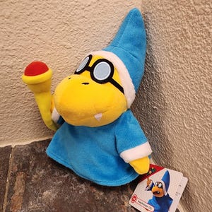 Pode incluir: Um peluche da personagem Magikoopa, com uma túnica azul com rebordo branco, um chapéu pontiagudo azul a condizer, óculos redondos de armação preta, um focinho amarelo e uma varinha amarela com ponta vermelha. Uma etiqueta Magikoopa (S) está presa.