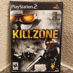 Może przedstawiać: Etui na grę wideo PlayStation 2 Killzone z czarno-białą sceną wojenną przedstawiającą żołnierzy w świecących pomarańczowych goglach. Tytuł Killzone jest wydrukowany dużymi żółtymi literami. Zawiera logo Sony i ocenę ESRB Mature 17+.