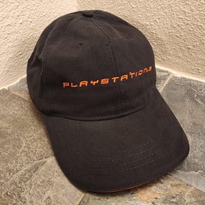 May include: A black baseball cap featuring the text PLAYSTATION 3 embroidered in orange thread across the front panels. The cap has a structured crown, a curved brim, and a small orange accent visible on the underside of the brim edge.