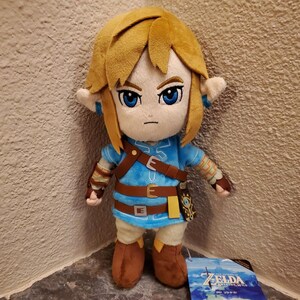May include: A plush toy of a character with blonde hair, pointed ears, and blue eyes. The figure wears a blue tunic with brown straps, a brown belt, and brown boots. A tag attached to the base displays the text The Legend of Zelda Breath of the Wild.