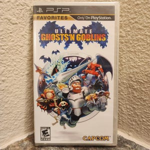 Peut inclure: Un boîtier de jeu vidéo pour PlayStation Portable intitulé Ultimate Ghosts 'n Goblins. La couverture représente un chevalier en armure argentée tenant une lance, entouré de monstres, squelettes et chauves-souris sur un fond circulaire.