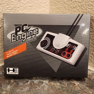 May include: A rectangular product box for the PC Engine mini game console. The box features a black and white horizontal striped pattern, the PC Engine mini logo, and an image of a white game controller with black and red buttons.