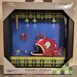 May include: A Pixel Frames shadow box art display featuring Mega Man 2 Lantern Fish. The scene shows a blue Mega Man character facing a large red angler fish against a blue background. The box measures 23x23 cm and includes official branding.