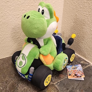 May include: A plush green Yoshi character sitting in a green and black toy go-kart. The kart features black wheels with yellow centres, a white stripe on the bonnet, and a blue seat. A Mario Kart World tag is attached to the side of the toy vehicle.