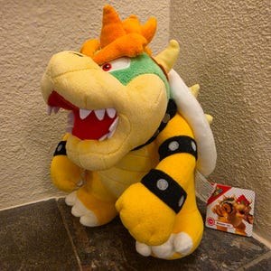 Puede incluir: Un peluche del personaje Bowser de Super Mario, con cuerpo amarillo, cabeza verde, pelo naranja y un caparazón blanco con púas. La figura tiene muñequeras negras con tachuelas plateadas, una boca roja con dientes blancos y una etiqueta.