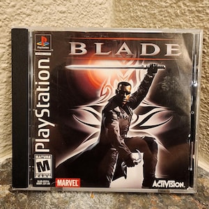 Op de afbeelding: Een PlayStation-videogamehoesje voor de titel Blade, met de figuur Blade die een zwaard boven het hoofd houdt. De hoes bevat de logo's van Marvel en Activision, een Mature ESRB-beoordeling en het PlayStation-logo op een zwarte achtergrond.