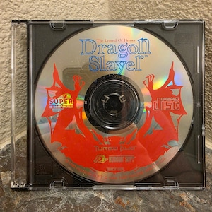 Peut inclure: Un disque de jeu vidéo Dragon Slayer: The Legend of Heroes pour le système Super CD-ROM2 dans un boîtier en plastique transparent. Le disque présente une surface argentée réfléchissante avec des silhouettes graphiques rouges et du texte bleu.