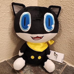 May include: A plush toy of a black cat character with large blue eyes, a yellow neck scarf, and white paws. The character has a white lower face, two small yellow buttons on the torso, and pointed ears with grey inner sections. A white tag reads Persona 5.