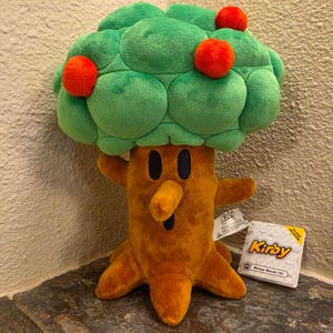 Puede incluir: Un peluche con forma de árbol que tiene un tronco marrón, una gran copa verde y tres frutos redondos de color naranja. El tronco presenta dos ojos ovalados negros y una nariz prominente. Una etiqueta blanca lateral muestra el texto Kirby.