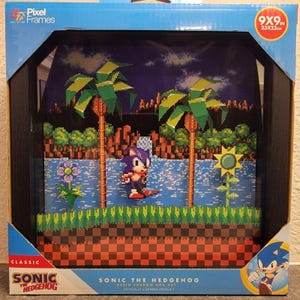 Op de afbeelding: Een Pixel Frames Sonic the Hedgehog schaduwdoos kunstwerk van 23x23cm. De zwart ingelijste doos bevat een gelaagde 3D pixel art scène van Sonic in een spelomgeving met palmbomen, een bloem, een zonnebloem en een schaakbordpatroon op de grond.