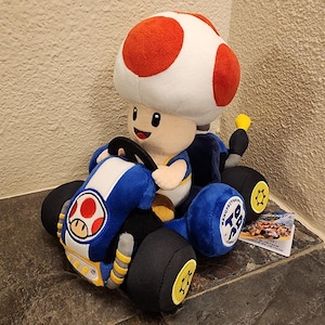 Puede incluir: Un peluche de Toad de Mario Kart sentado en un kart de carreras de juguete azul y blanco. El kart cuenta con ruedas negras con detalles amarillos, un pequeño volante y un emblema de Toad en el capó delantero. Tiene una etiqueta adjunta.