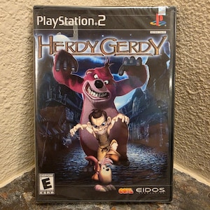 May include: A factory-sealed PlayStation 2 video game case for Herdy Gerdy. The cover art features a large pink creature, a human character, and a small animal on a cobblestone street. The PlayStation 2 logo, Eidos logo, and ESRB E rating are visible.