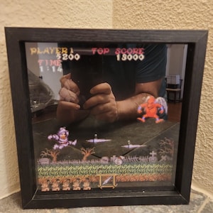 Puede incluir: Una caja de sombra cuadrada con marco negro que muestra un diorama 3D multicapa de una escena de videojuego retro. La escena presenta un personaje pixelado, una criatura roja voladora y un paisaje de cementerio con el texto PLAYER 1, 5200, TOP SCORE, 18000, TIME y 1:14.