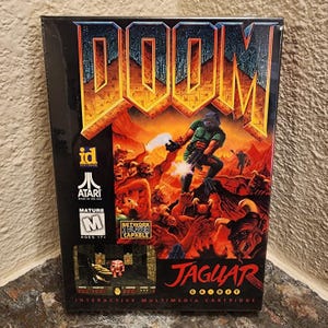 Pode incluir: Uma caixa de jogo de vídeo selada para Doom na Atari Jaguar. A capa apresenta um soldado com armadura verde a lutar contra demónios numa paisagem ardente. Estão presentes logótipos da id Software e Atari, uma classificação Mature M e um ecrã.
