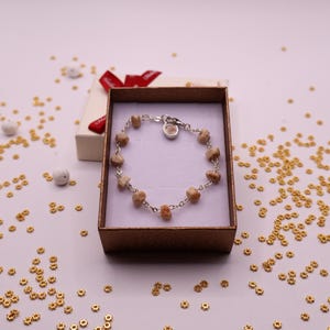 May include: A silver-toned chain bracelet featuring small, textured, light brown spherical beads spaced evenly along the links. The bracelet is displayed inside a rectangular brown cardboard gift box with a white interior and a red ribbon.