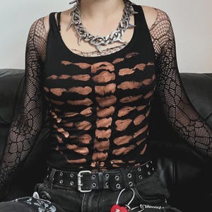 Op de afbeelding: Een zwart hemdje met een bruine print van een skelet-borstkas, gedragen over zwarte visnet-mouwen. De outfit bevat een zilverkleurige grove schakelketting en een zwarte leren riem met zilveren metalen oogjes en een rechthoekige gesp.