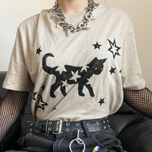 Könnte beinhalten: Ein beiges Kurzarm-T-Shirt mit einem schwarzen Grafikdruck einer Katze, die mit Sternenmustern bedeckt ist und von weiteren schwarzen Sternen umgeben wird. Das T-Shirt wird mit einer dicken silbernen Gliederkette, schwarzen Netzärmeln und einem schwarzen Gürtel kombiniert.