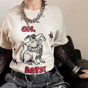 Puede incluir: Una camiseta beige de manga corta con un gráfico en negro y rojo de una rata con gorro y cuello de bufón. El texto "OH, RATS!" está impreso en rojo sobre y bajo la ilustración, rodeado de pequeñas formas de estrellas negras y rojas.
