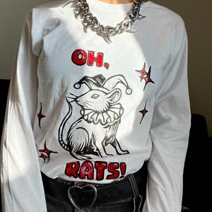 Puede incluir: Una camiseta blanca de manga larga con un gráfico en negro y rojo de una rata con gorro y cuello de bufón. El texto "OH. RATS!" está impreso en rojo sobre y bajo la ilustración, rodeado de pequeñas formas similares a estrellas.