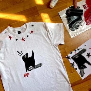 Puede incluir: Una camiseta blanca de manga corta con un gráfico negro de un gato aullando, detalles de estrellas rojas y negras alrededor del cuello y una pequeña firma roja debajo del gato. El diseño está impreso con una estética texturizada de tinta.