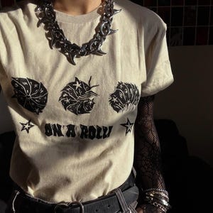 Peut inclure: Un t-shirt beige à manches courtes orné de trois illustrations graphiques noires représentant des cloportes et du texte "ON A ROLL" avec des étoiles. Le t-shirt est porté avec un collier à chaîne métallique argentée aux maillons pointus.